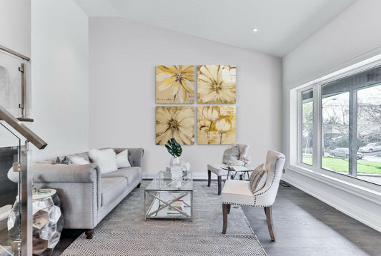 Four-panel yellow daisy and flower glass wall art displayed above a gray sofa in a bright living room with large windows.