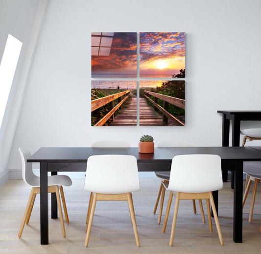 Four-panel glass wall art depicting a wooden boardwalk leading to a vibrant sunset over the ocean, displayed above a dining table.