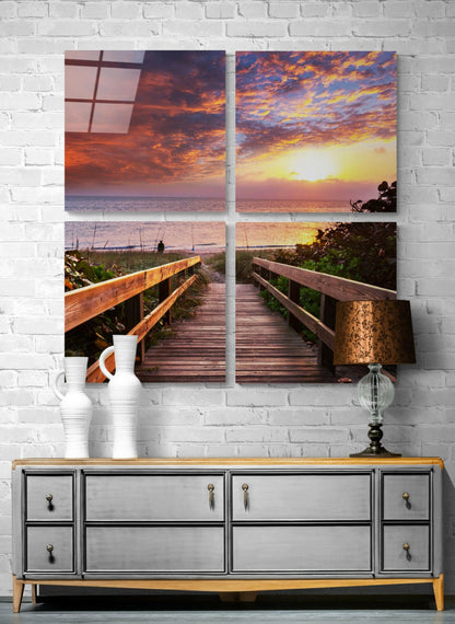 Four-panel glass wall art displaying a wooden boardwalk leading to a vibrant sunset ocean view with dramatic orange and purple sky.