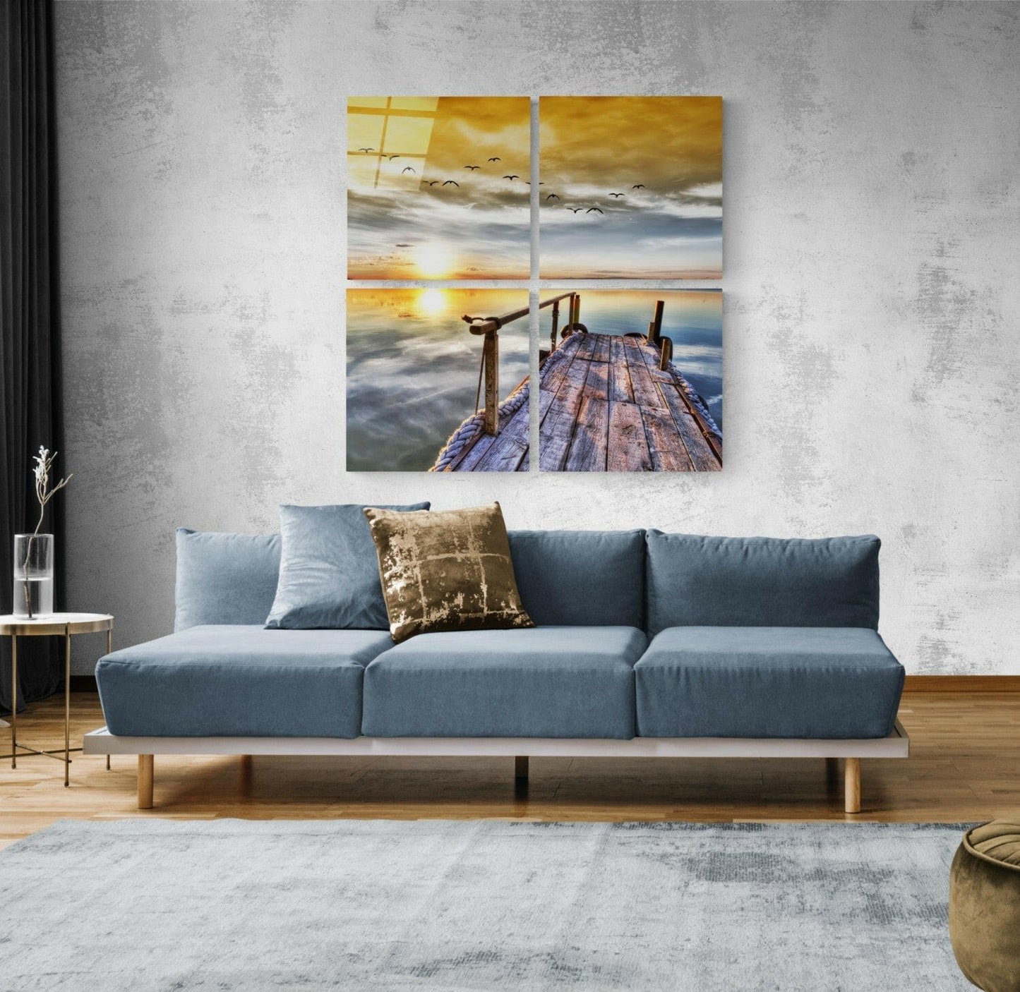 Four-panel glass wall art depicting a wooden pier at sunset with golden sky, birds in flight, and calm water, displayed above a blue sofa in a modern living room.
