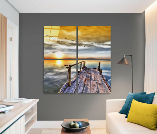 Four-panel glass wall art depicting a wooden pier at sunset with golden sky, birds in flight, and calm water, displayed above a cream sofa with teal and yellow pillows in a modern living room.