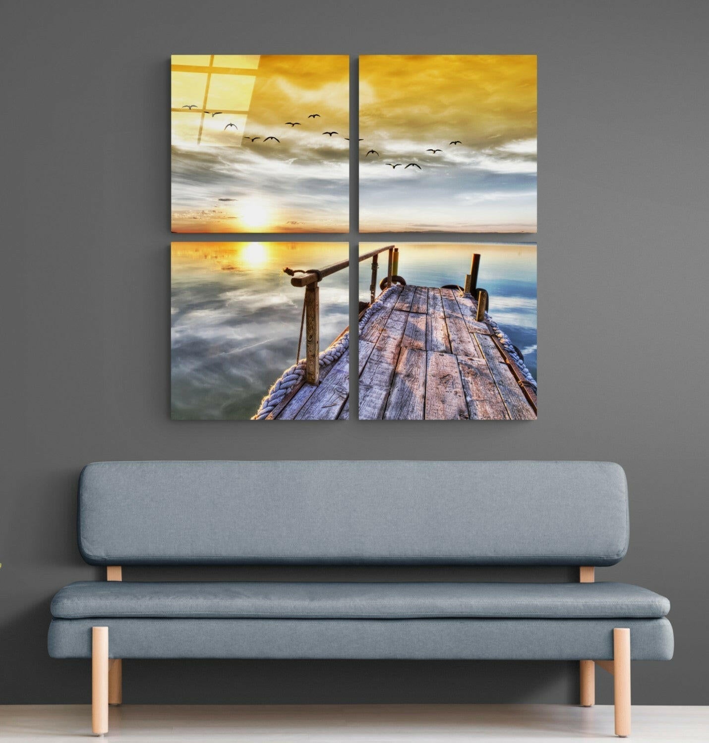Four-panel glass wall art displaying wooden pier extending into water at sunset with golden sky and flying birds, hung above gray upholstered bench.