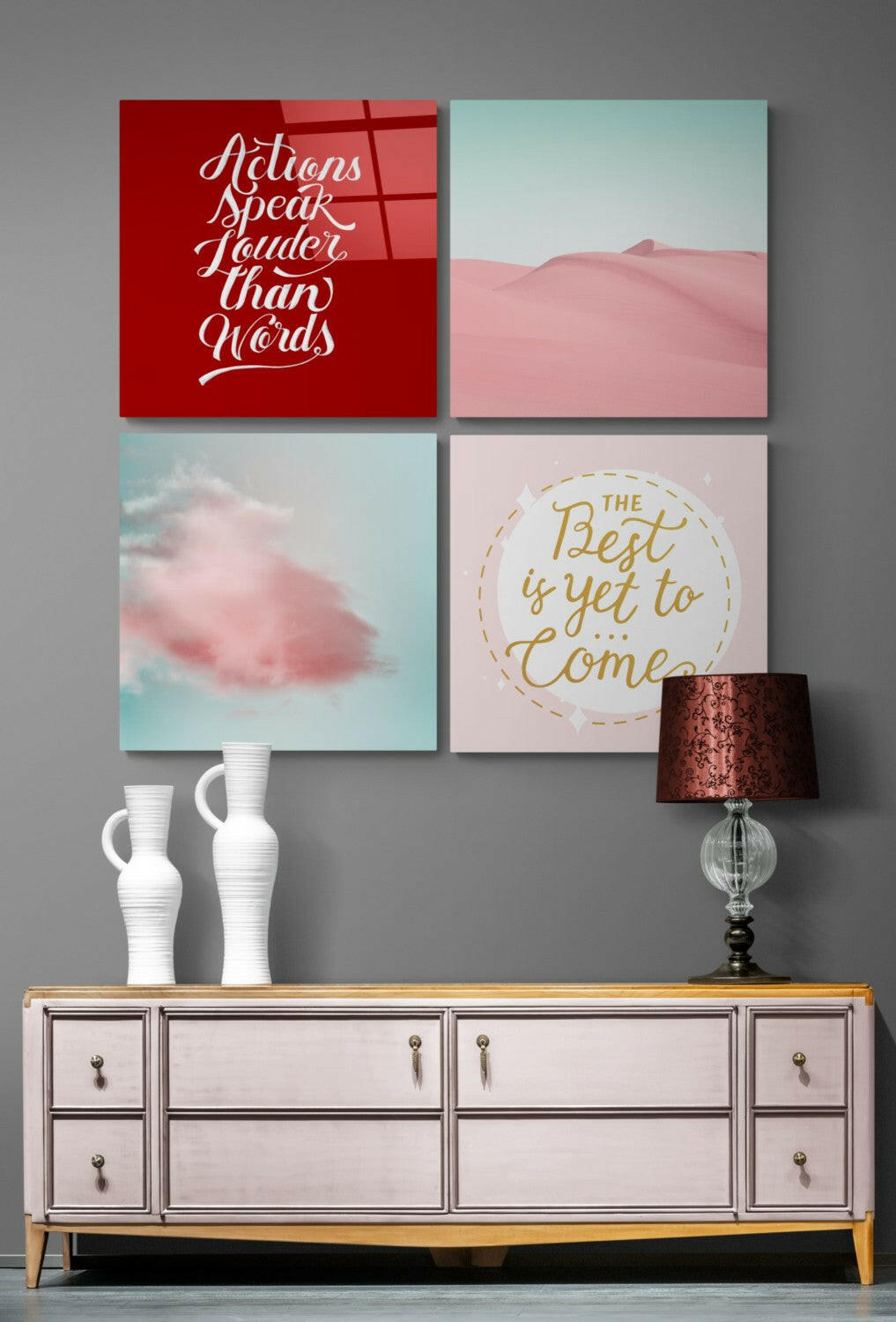 Four glass wall art panels displayed above a white dresser with decorative vases and a lamp, featuring motivational text and pink desert landscapes.