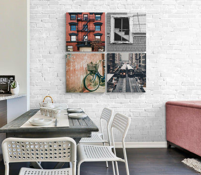Four glass wall art panels displaying urban scenes including a red brick building, gray house window, vintage bicycle, and city street with train tracks, installed above a dining table.