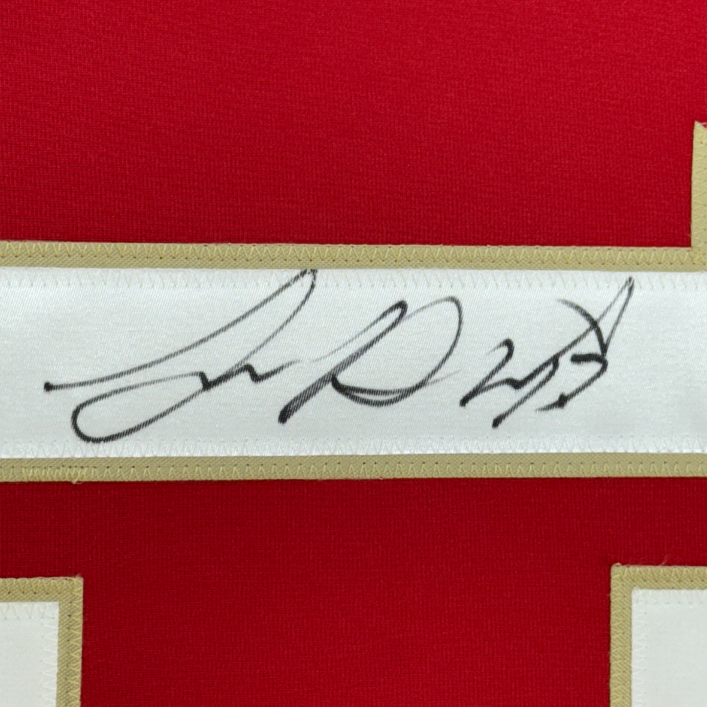 Close-up of handwritten signature in black ink on white fabric strip bordered by gold trim and red jersey material.