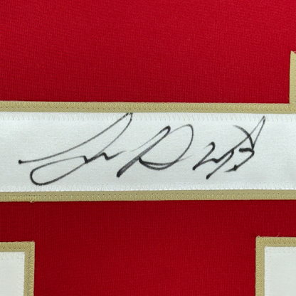 Close-up of handwritten signature in black ink on white fabric strip bordered by gold trim and red jersey material.