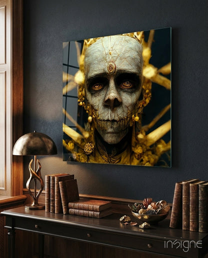 Glass wall art depicting a skeletal face with golden ornaments and jewelry displayed above a dark desk with books and lamp.
