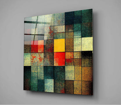 Angled view of a square glass wall art panel with a grid of textured squares in yellow, red, green, teal, brown, and cream colors against a gray background.
