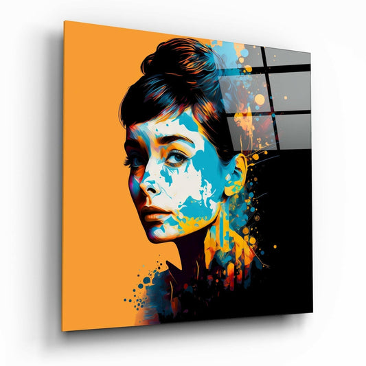 Angled perspective of glass wall art displaying a woman's profile portrait with orange background, blue and yellow paint splatters, and black geometric grid pattern on right side.