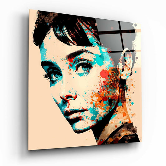 Glass wall art displayed at an angle showing a colorful portrait with teal, orange, and black splatter effects on a cream background.