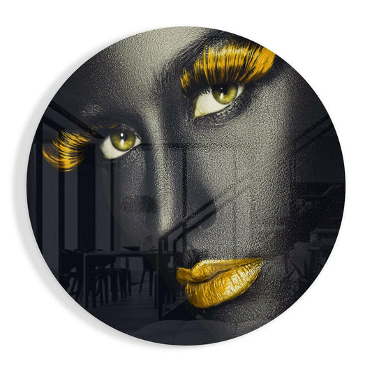 Circular glass wall art featuring a gray-faced figure with striking yellow-gold eyelashes, eyes, and lips against a dark background.