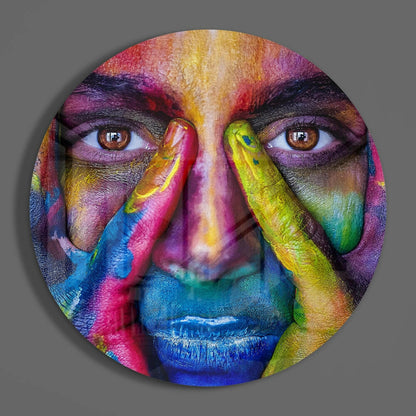 Circular glass wall art featuring a colorful portrait with rainbow-painted face, hands, and vibrant multicolored pigments covering the entire surface.
