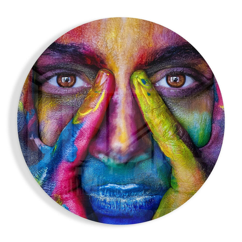 Circular glass wall art featuring a colorful portrait with rainbow-painted face, hands near face, and vibrant multicolored pigments covering skin and features.