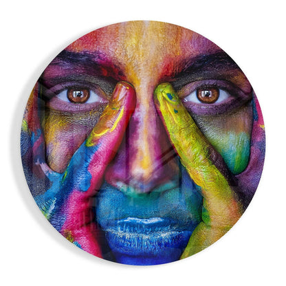 Circular glass wall art featuring a colorful portrait with rainbow-painted face, hands near face, and vibrant multicolored pigments covering skin and features.