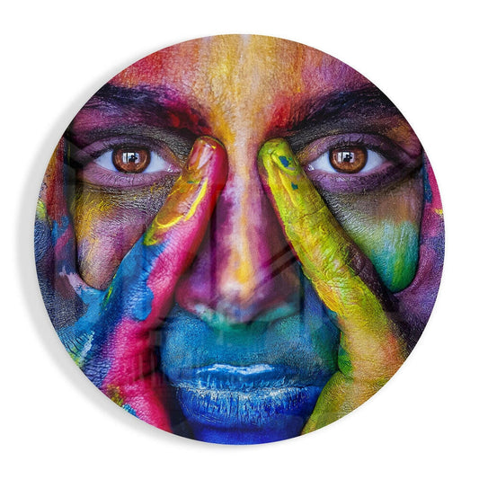 Circular glass wall art featuring a colorful portrait with rainbow-painted face, hands near face, and vibrant multicolored pigments covering skin and features.