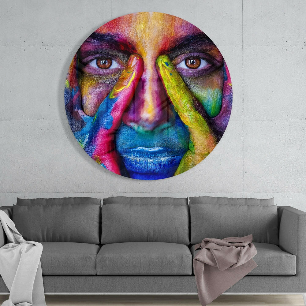 Vibrant circular glass wall art featuring a colorful face with rainbow-painted features and hands, displayed above a gray sofa.