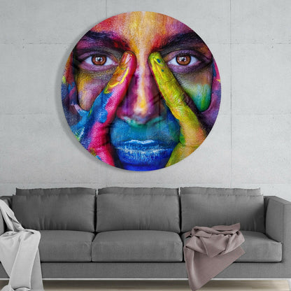 Vibrant circular glass wall art featuring a colorful face with rainbow-painted features and hands, displayed above a gray sofa.