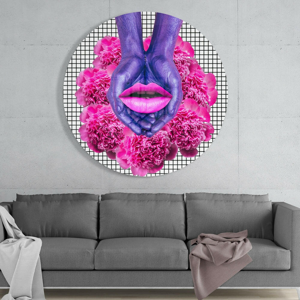 Circular glass wall art featuring purple hands forming a face with bright pink lips and carnation flowers, displayed above a gray sofa.