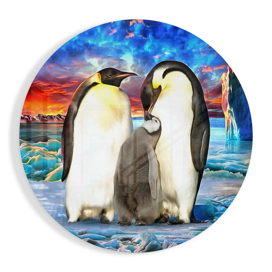 Round glass wall art featuring two adult emperor penguins with a chick between them against an icy Antarctic landscape with colorful sunset sky.