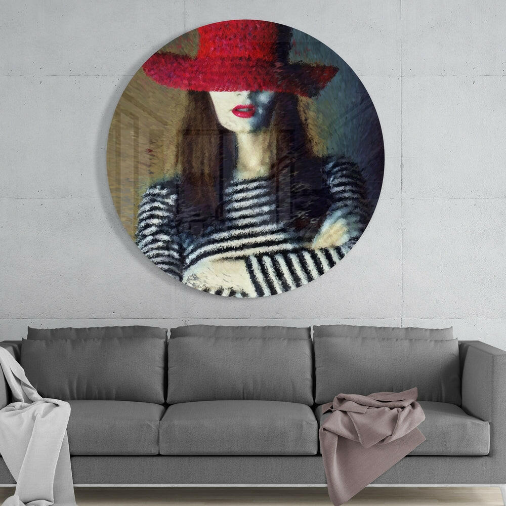 Circular glass wall art depicting a woman wearing a red hat and striped clothing, mounted above a gray sofa in an interior setting.