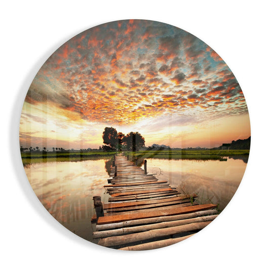 Wooden log bridge stretching across still rice paddies under a dramatic sunset sky with orange and gold clouds.