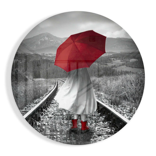 Person holding red umbrella and wearing white dress and red boots standing on railroad tracks in black and white mountain landscape.