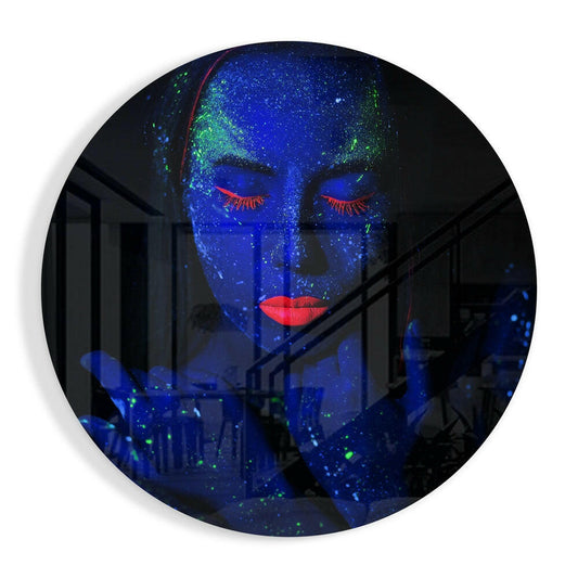 Round glass wall art displaying a woman's face with closed eyes in blue, green speckled paint, red eyelashes and lips, against an interior background.