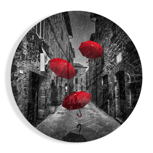 Circular glass wall art depicting three red umbrellas floating above a rainy, narrow European stone street in black and white.
