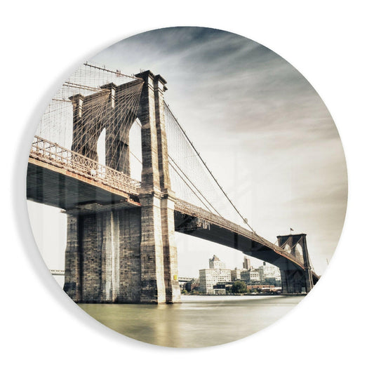 The Brooklyn Bridge spans across water with stone towers and cable supports visible, printed on a circular glass panel with sepia tones and cloudy sky background.