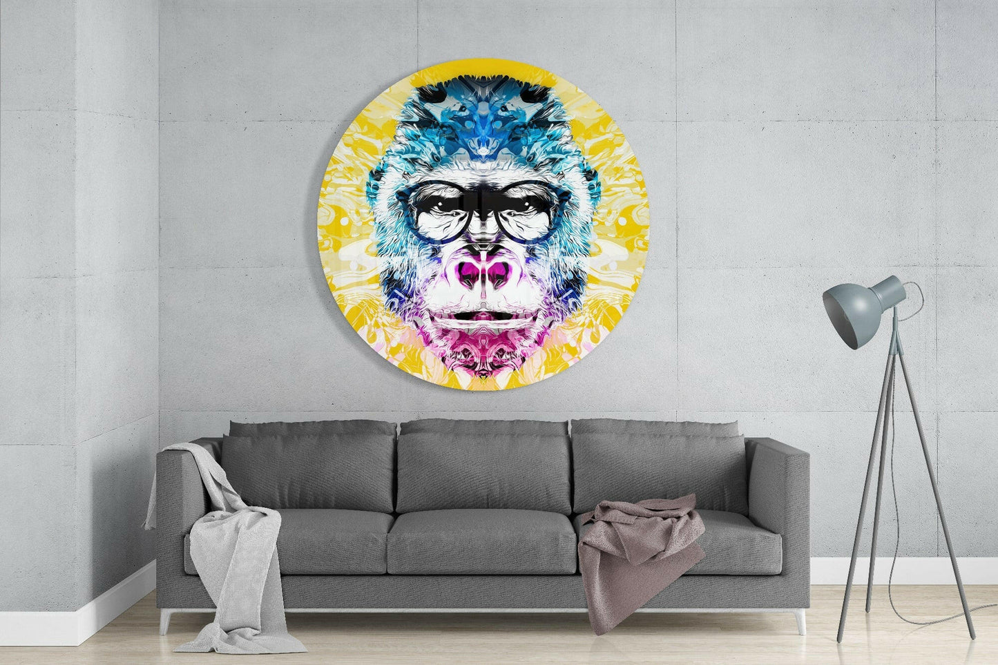 Circular glass wall art depicting a colorful gorilla with blue fur, pink mouth, black sunglasses, and yellow radiating background, mounted above gray sofa in modern living room.