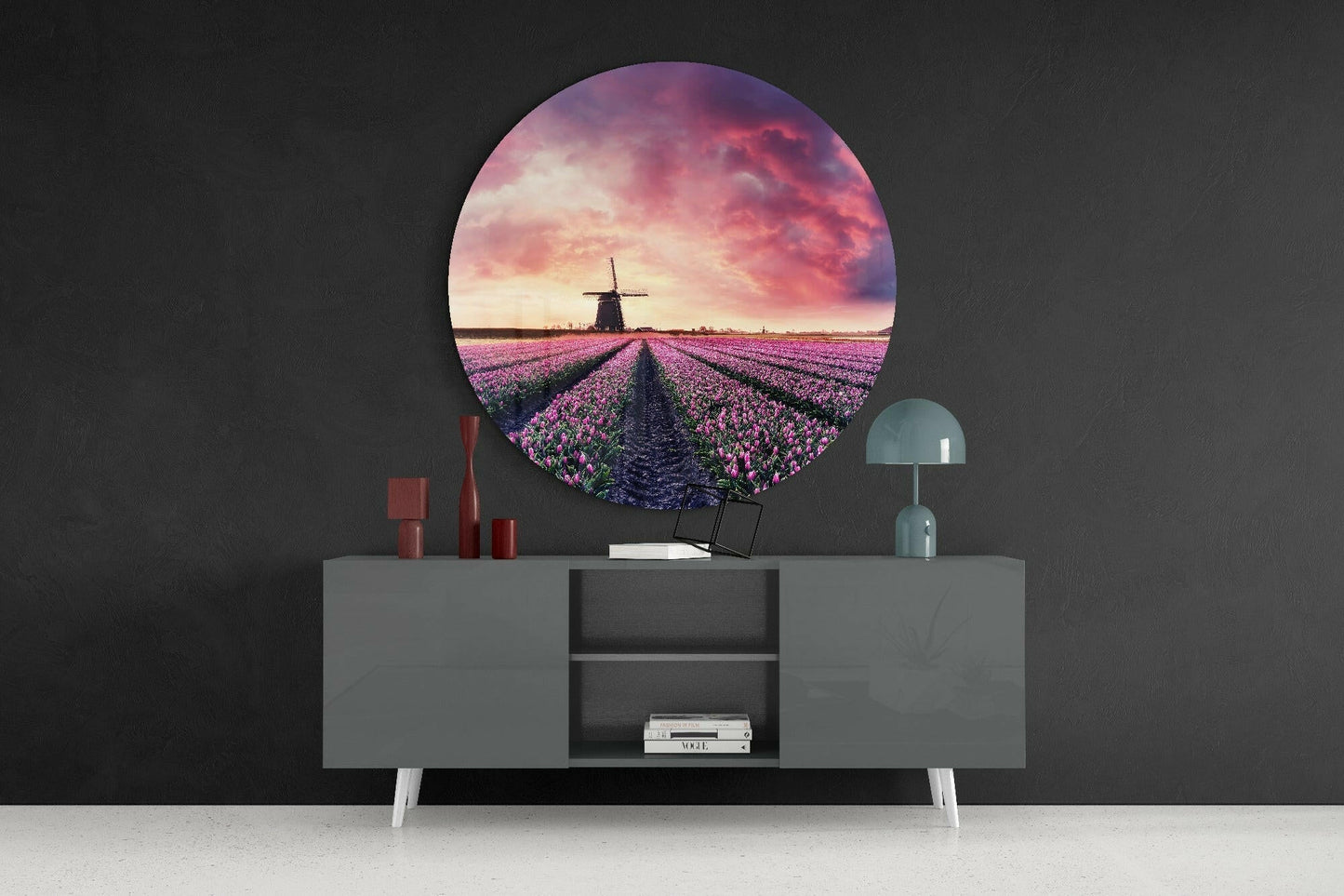 Circular glass wall art displaying purple tulip field with windmill at sunset, mounted above grey sideboard with red candles and teal lamp.