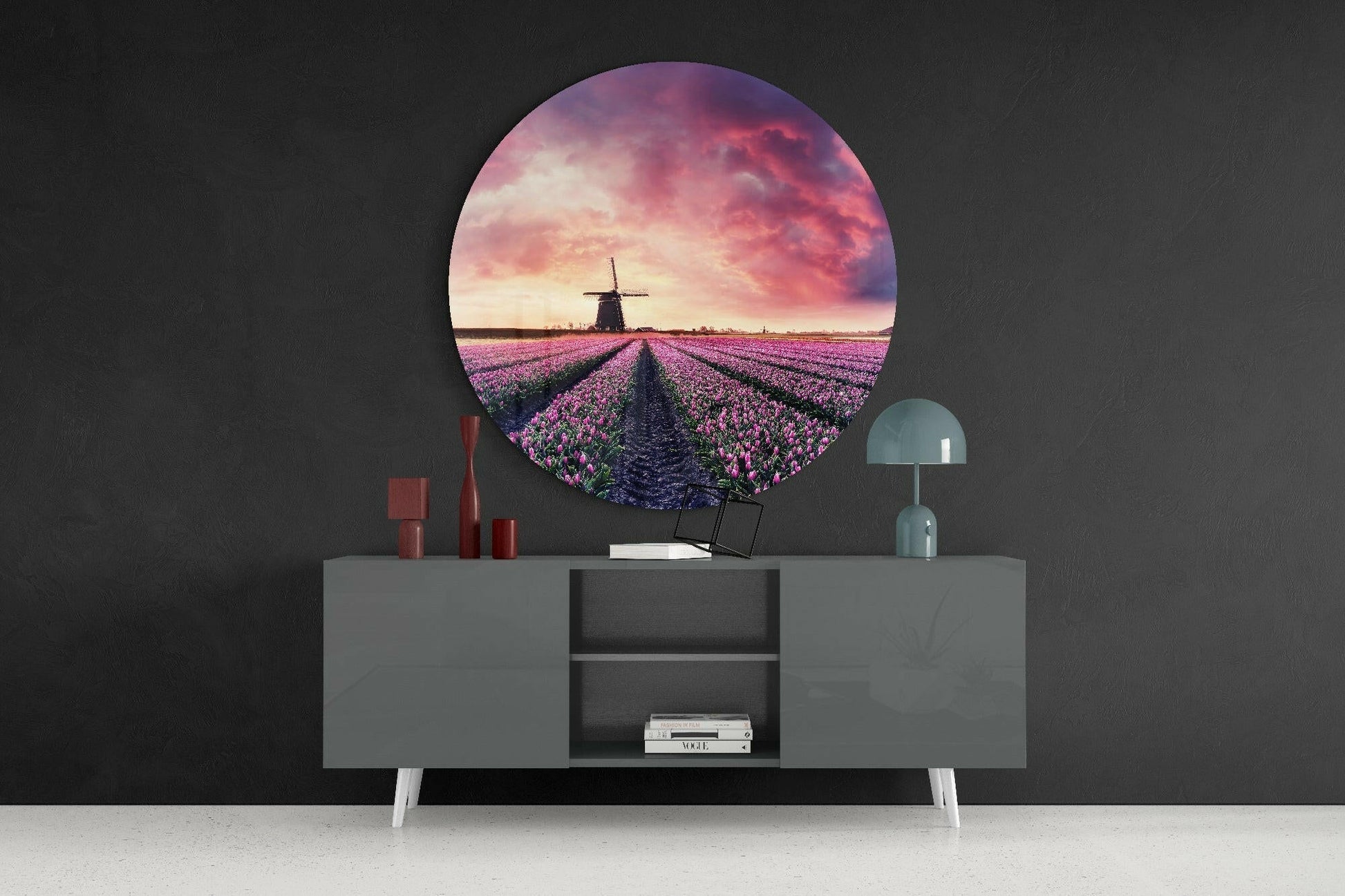 Circular glass wall art displaying purple tulip field with windmill at sunset, mounted above grey sideboard with red candles and teal lamp.
