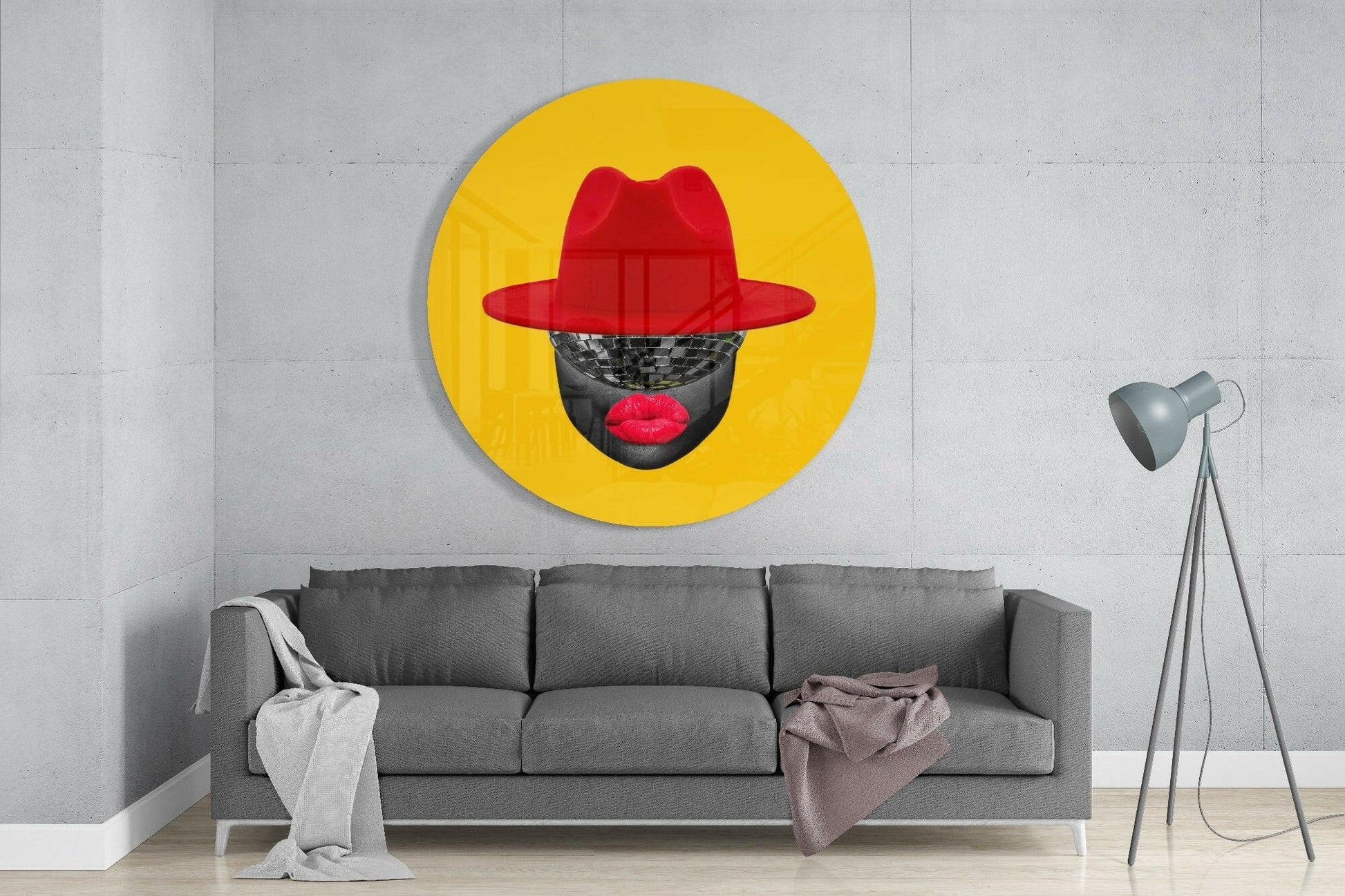Round yellow glass wall art with red hat and red lips on grayscale face, displayed above gray sofa in modern interior.