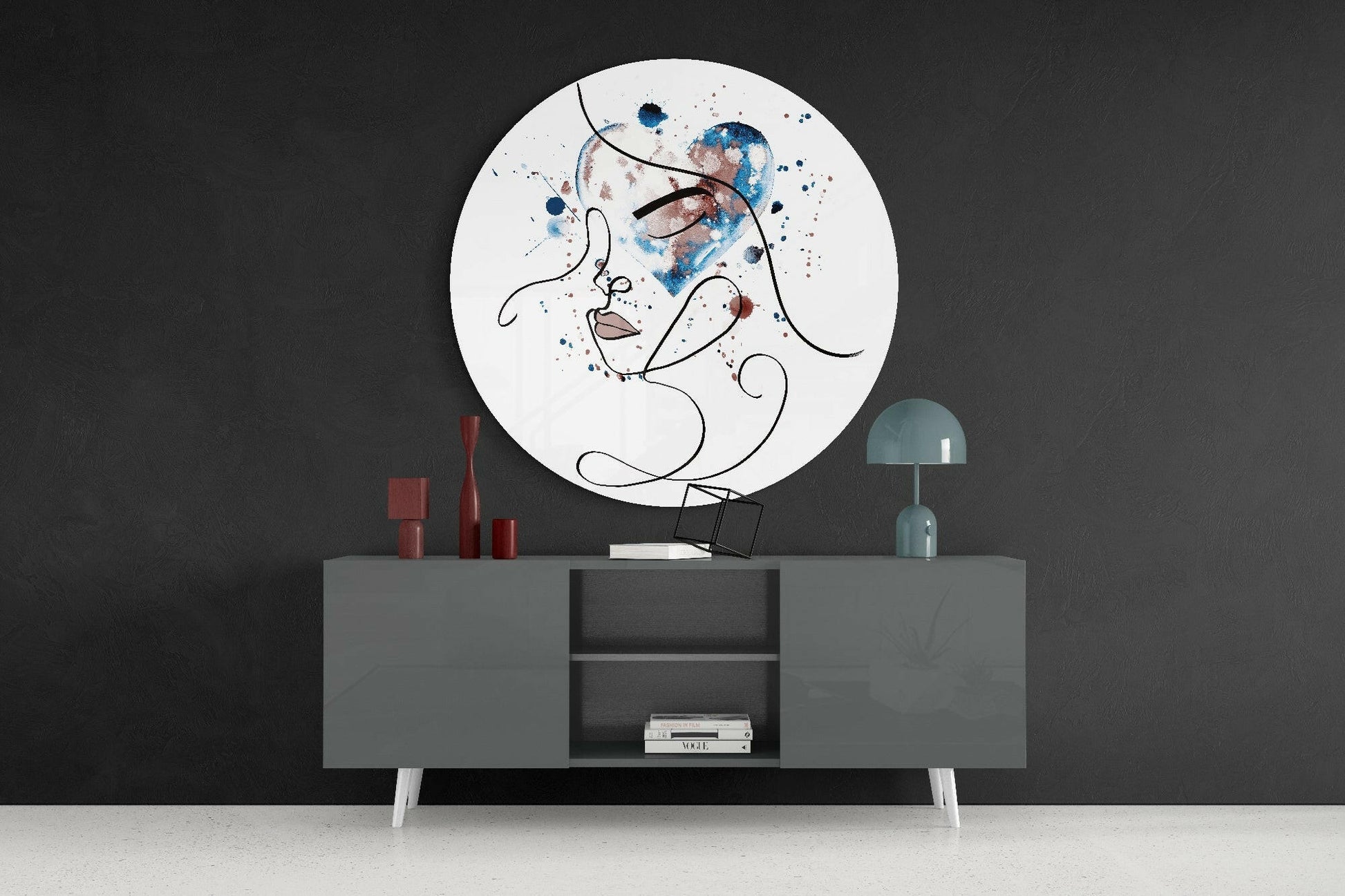 Circular glass wall art displaying abstract line-drawn face with blue and brown watercolor splashes, mounted above gray console table with red candles and teal lamp.