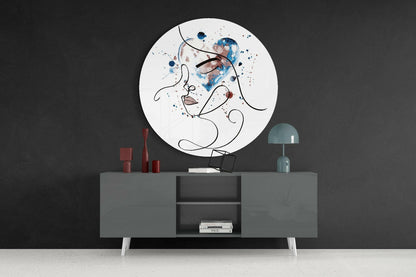 Circular glass wall art displaying abstract line-drawn face with blue and brown watercolor splashes, mounted above gray console table with red candles and teal lamp.