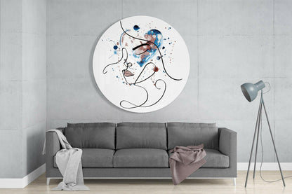 Circular white glass wall art with abstract line-drawn face and blue, brown watercolor splashes, displayed above gray sofa in modern living room.