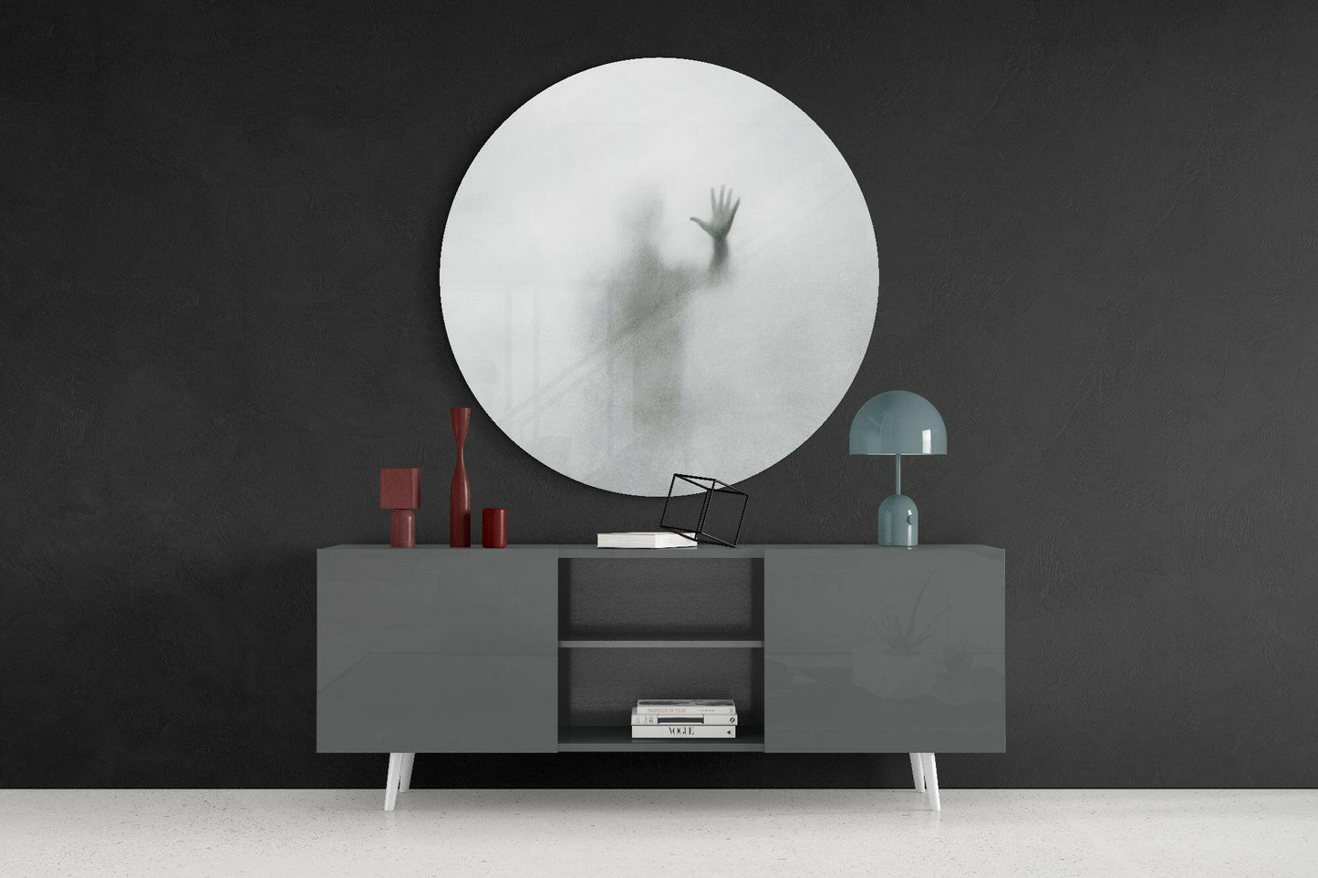 Large circular frosted glass wall mirror with blurred hand silhouette mounted above grey sideboard with red candles and teal lamp.