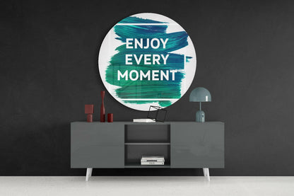 Circular glass wall art with teal brushstroke design and white "Enjoy Every Moment" text displayed above gray sideboard with decorative items.