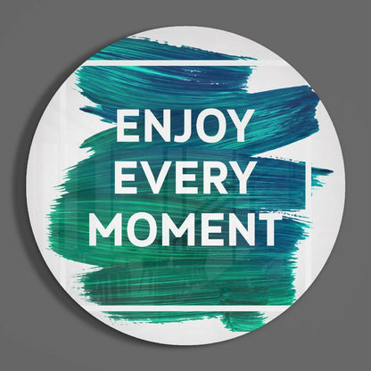 Round white glass wall art with teal and blue brushstroke design and white "ENJOY EVERY MOMENT" text, shown straight-on against gray background.