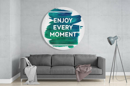 Round white glass wall art with teal and green brushstroke design displaying "ENJOY EVERY MOMENT" text, mounted above gray sofa in modern living room.