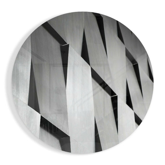 Circular glass wall art featuring overlapping silver and gray angular slats creating a three-dimensional geometric pattern with sharp shadows.