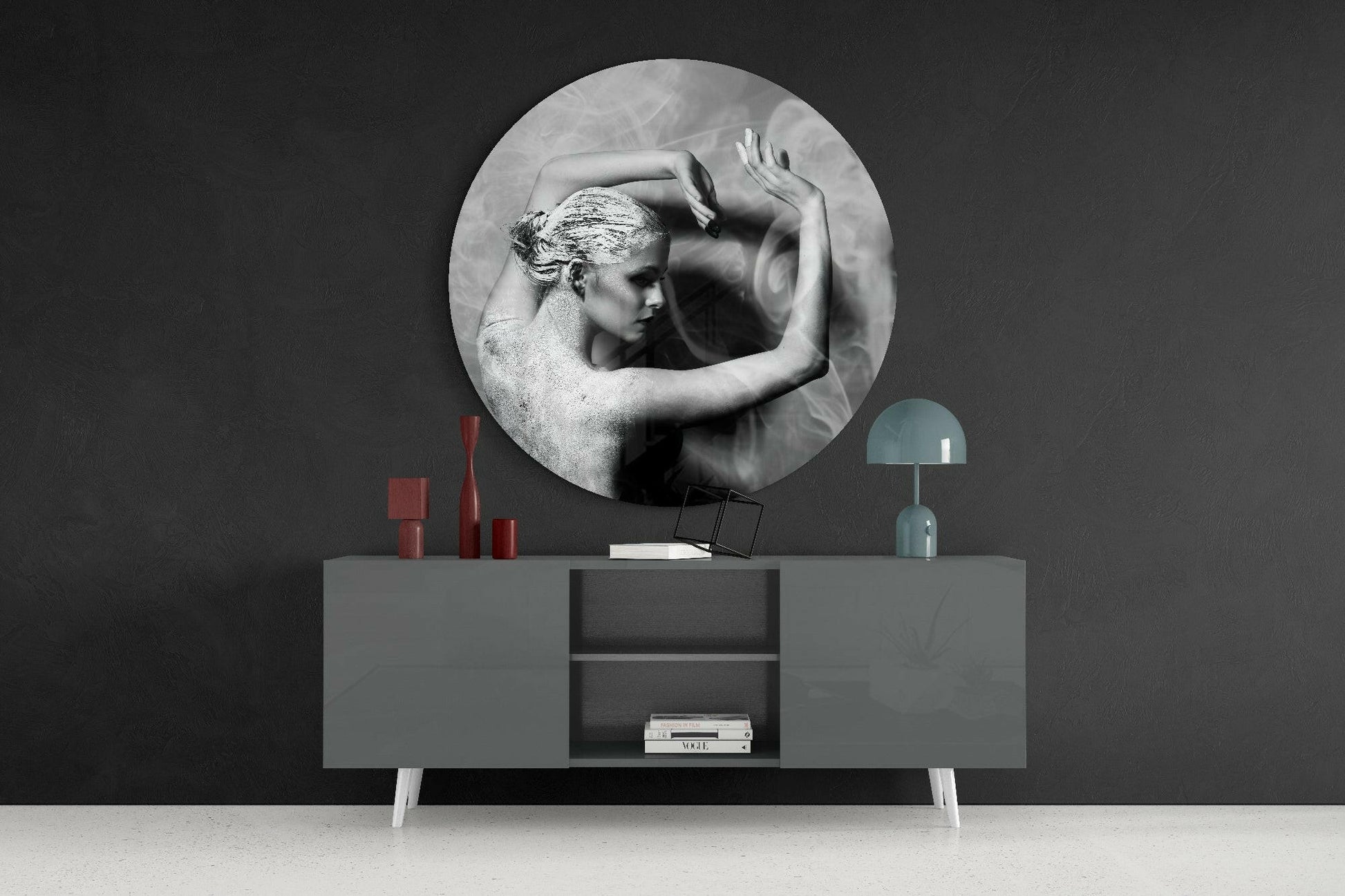 Circular black and white glass wall art depicting a classical female figure mounted above a gray sideboard with red decorative objects and a teal lamp.