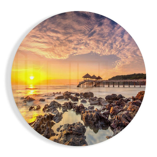 Sunset over rocky coastline with wooden pier pavilions reflected in calm water, displayed on circular glass wall art.
