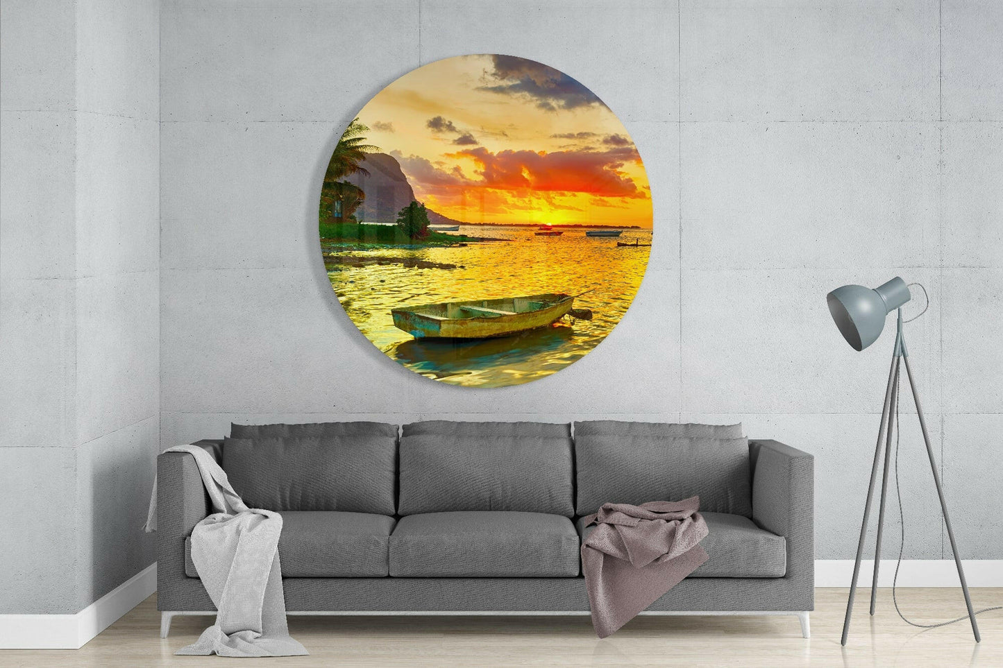 Circular glass wall art displaying a golden sunset over water with a moored fishing boat, mounted above a gray sofa in a minimalist interior.