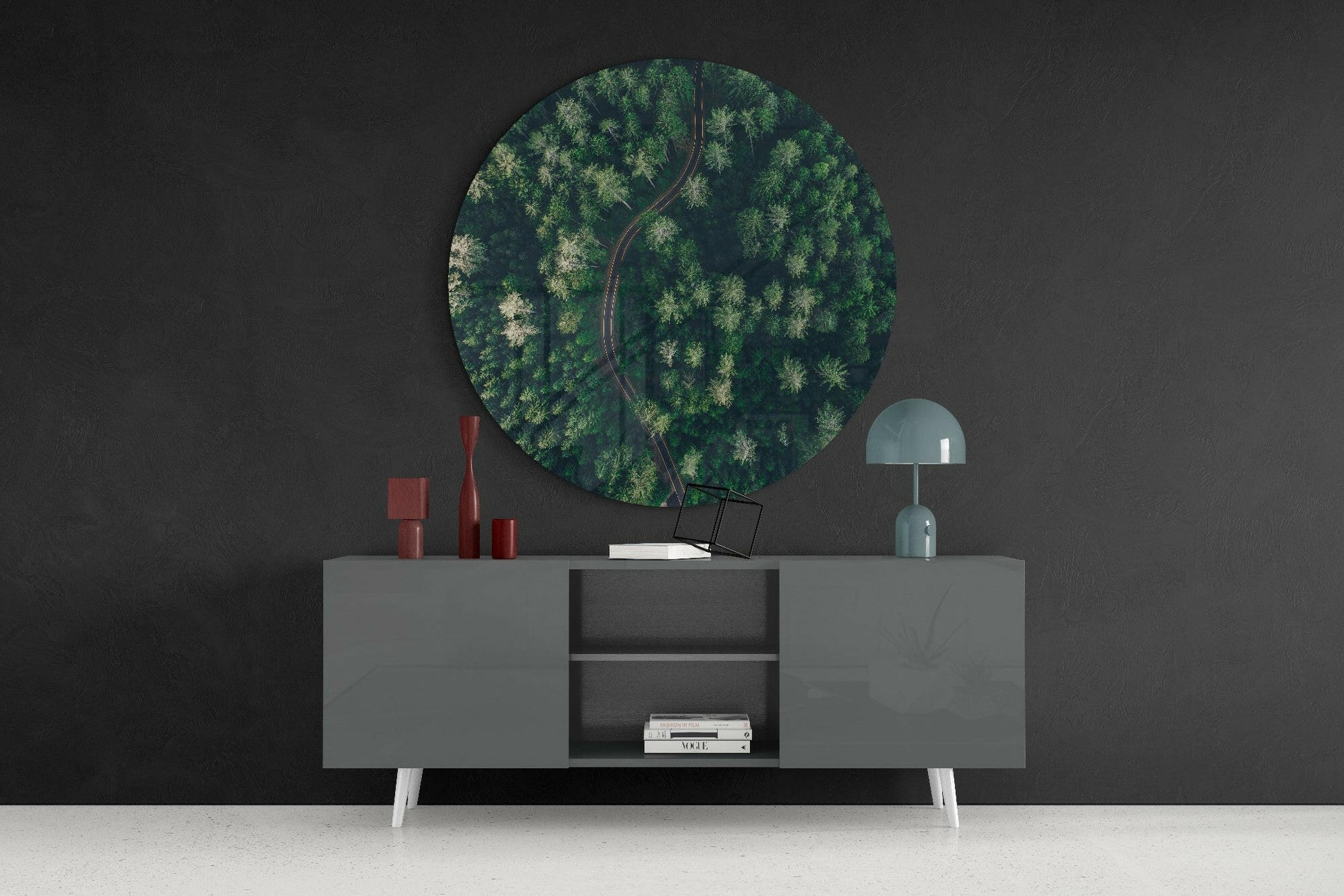 Circular aerial forest glass wall art with road displayed above gray sideboard with white legs against dark wall.