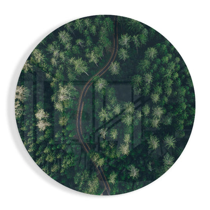 Aerial view of a winding red road cutting through a dense green forest with scattered trees, printed on circular glass wall art.