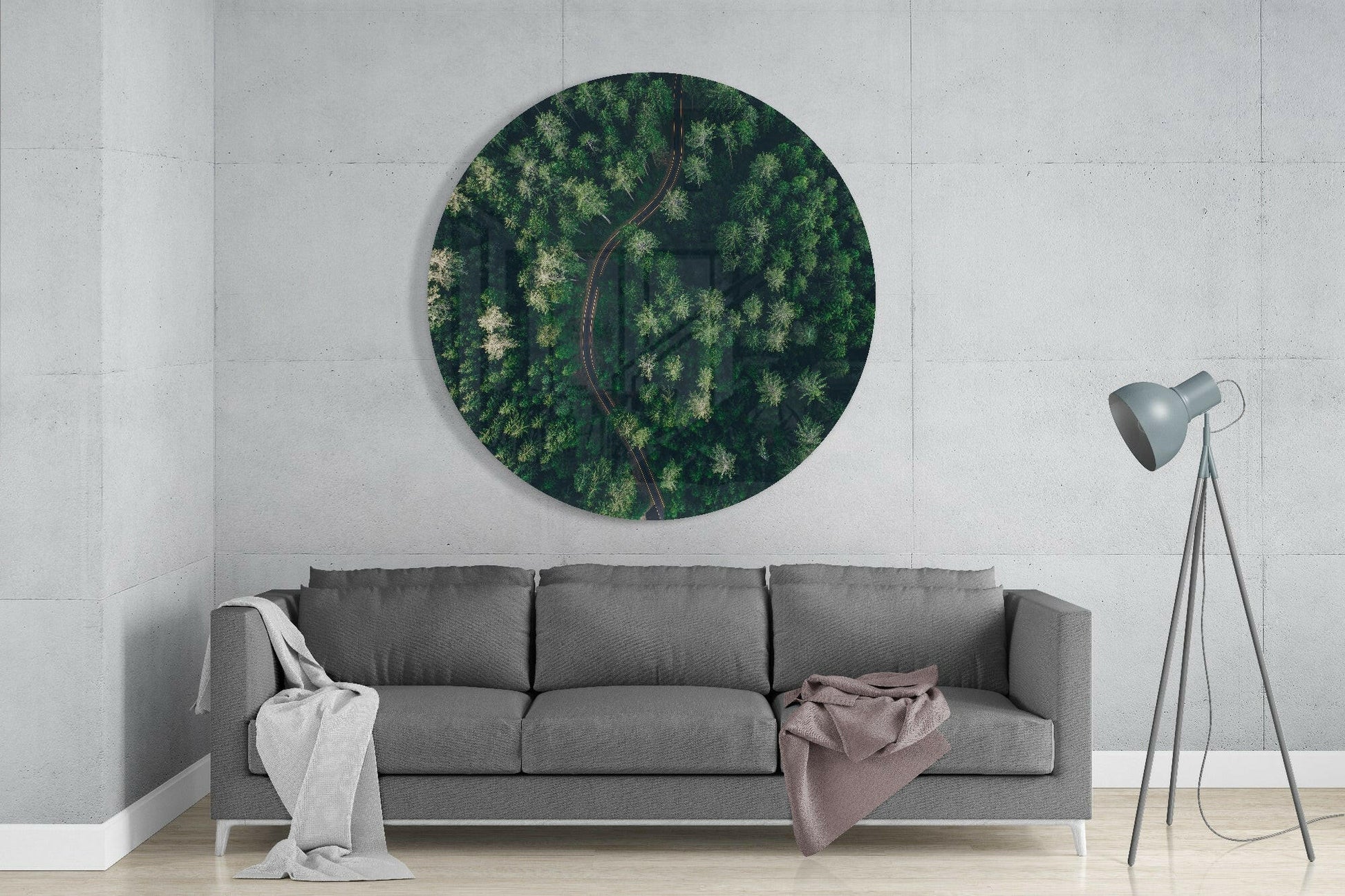 Gray sofa beneath circular glass wall art showing aerial forest view with winding road, concrete wall background.