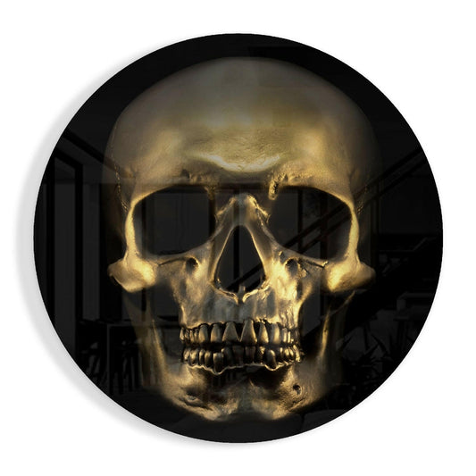 Front-facing bronze gold skull with detailed bone structure and teeth against dark background, mounted in circular glass frame.