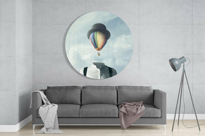 Circular glass wall art mounted above gray sofa displays surreal image of figure with bowler hat and hot air balloon replacing head against cloudy sky.