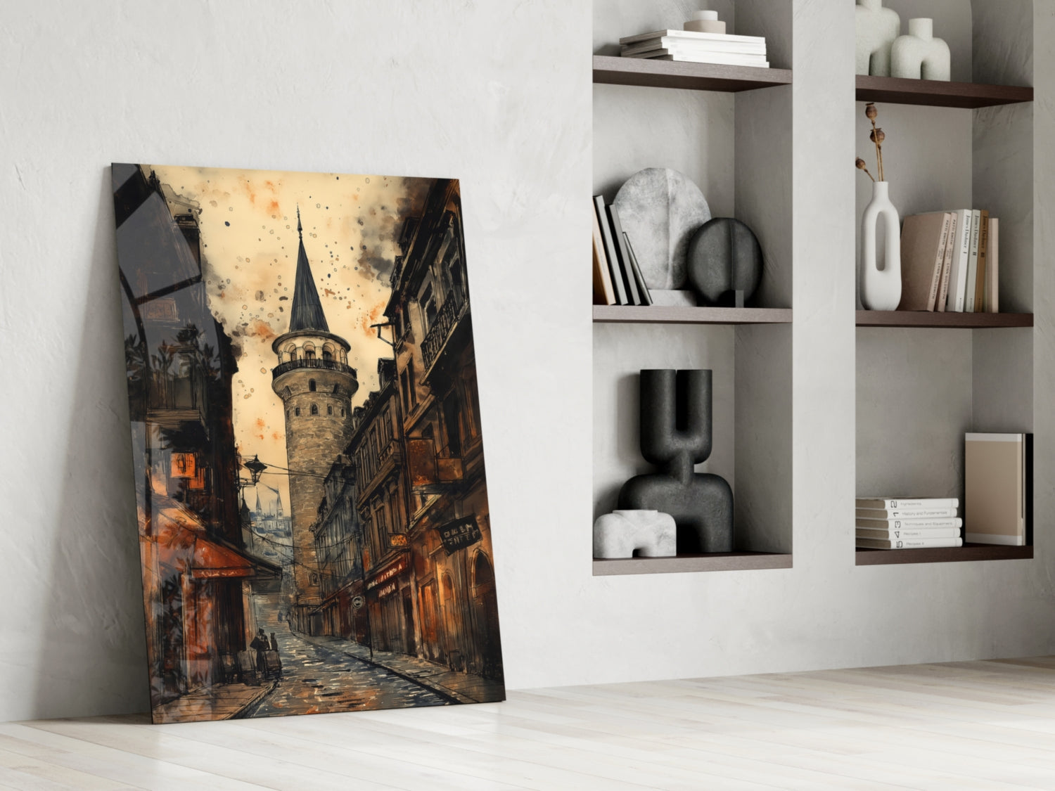Glass wall art leaning against white wall displays warm-toned street scene with historic tower, alongside styled shelving unit with decorative objects and books.